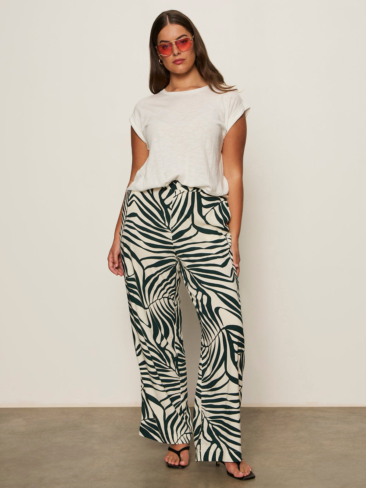 A woman with long brown hair and a relaxed pose stands against a plain wall wearing Sanctuary Clothing's the soft trouser green oasis extended sizing, a white t-shirt, black sandals, and pink sunglasses.
