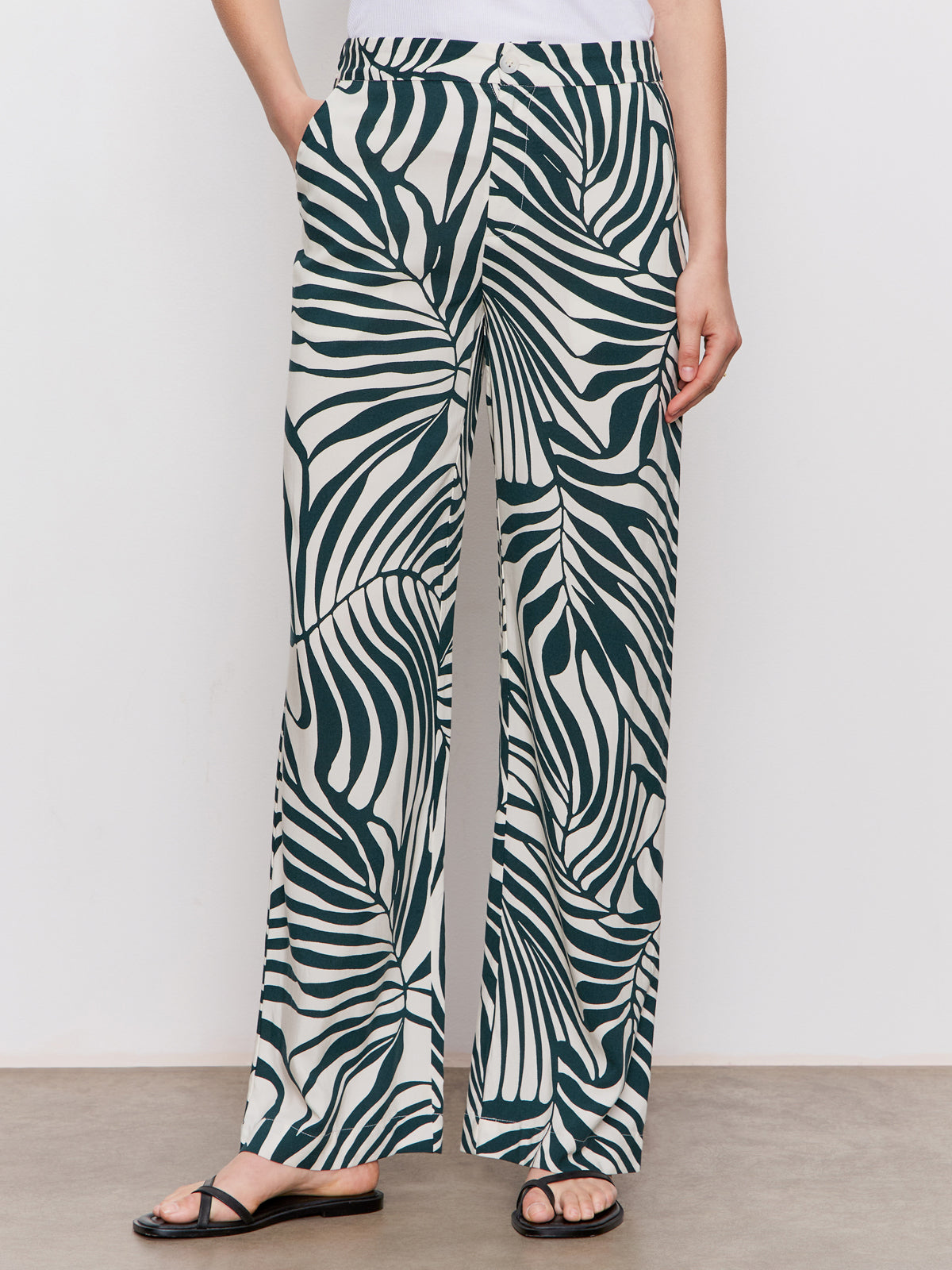 A person wearing Sanctuary Clothing's "the soft trouser green oasis," high-waisted wide-leg pants in a green abstract leaf print, paired with a white top and black sandals, stands against a plain background.