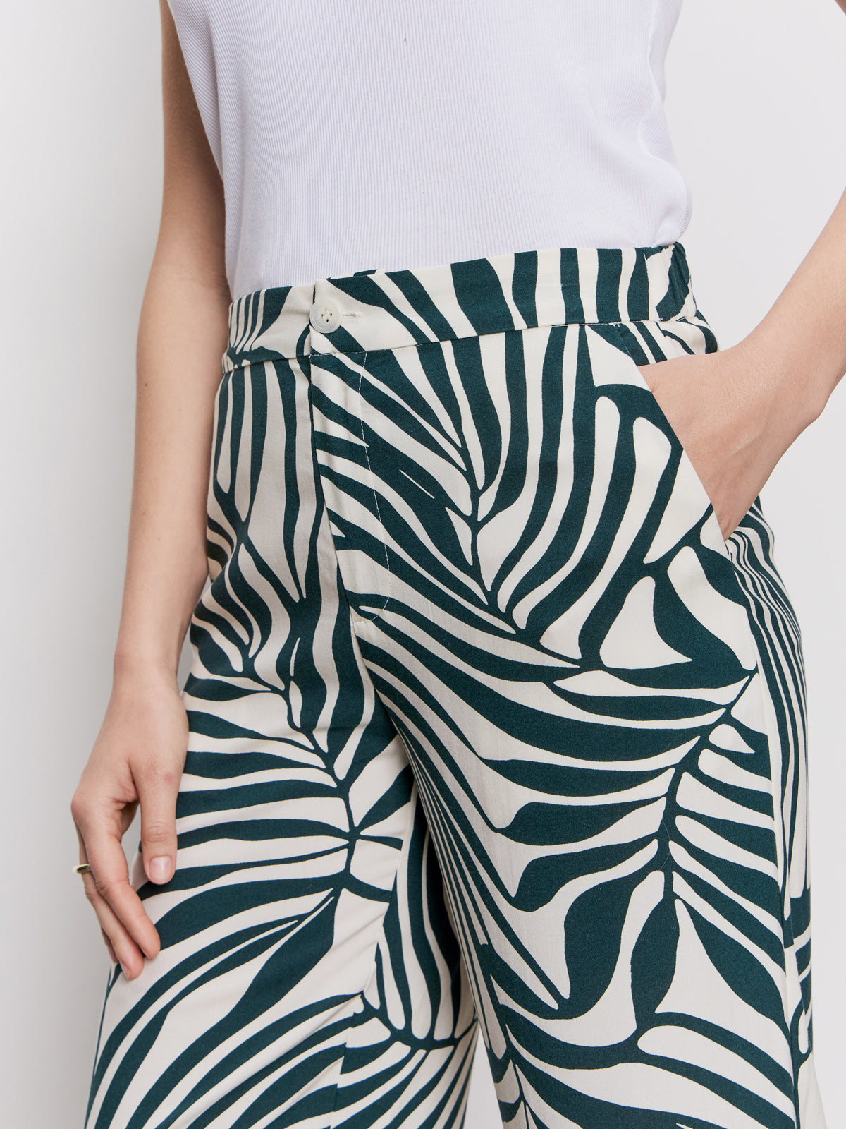 A person wears the Sanctuary Clothing "the soft trouser green oasis," featuring a bold green and white leaf print, paired with a sleeveless white top. One hand is in the pocket, and part of the arm and torso are shown.