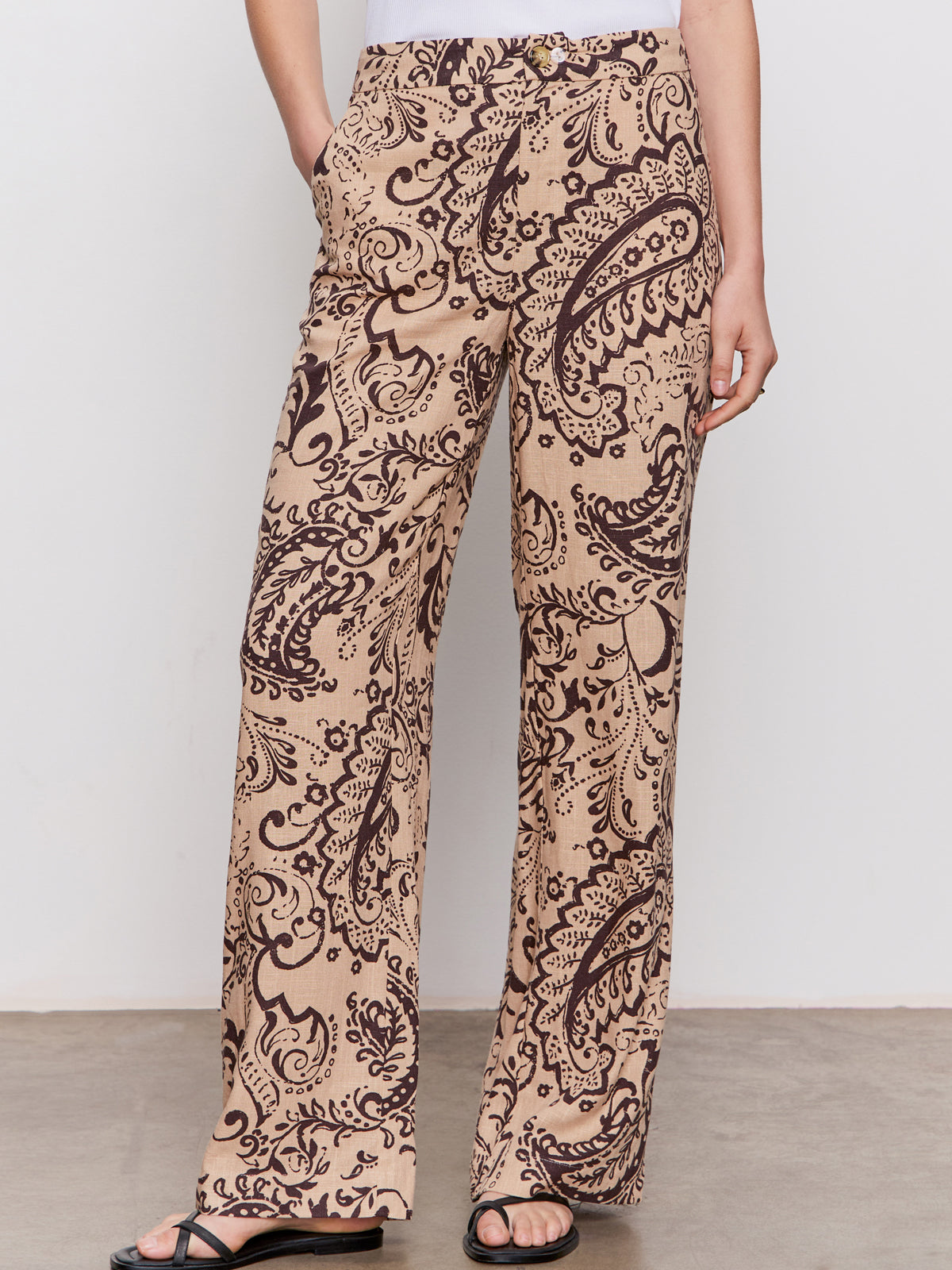 A person wears the soft trouser desert bandana by Sanctuary Clothing—beige wide-leg pants with a black paisley print—paired with a white top and black sandals, standing against a plain background.
