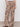 A person wears the soft trouser desert bandana by Sanctuary Clothing�beige wide-leg pants with a black paisley print�paired with a white top and black sandals, standing against a plain background.