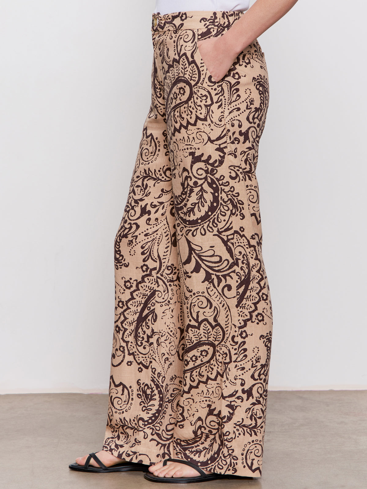 A person wearing Sanctuary Clothing’s the soft trouser desert bandana—high-waisted, wide-leg pants with a beige base and bold brown paisley print—stands sideways in black sandals against a plain background.