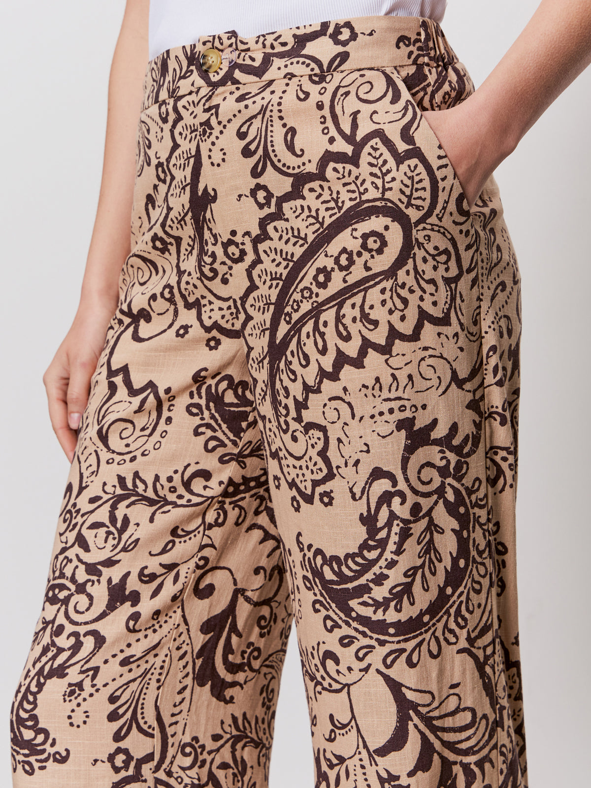 A person wearing Sanctuary Clothing's "the soft trouser desert bandana"—beige pants with a dark brown paisley and floral print—stands with one hand in their pocket, showing only the lower torso and upper legs.