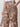 A person wearing Sanctuary Clothing's "the soft trouser desert bandana"�beige pants with a dark brown paisley and floral print�stands with one hand in their pocket, showing only the lower torso and upper legs.