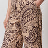 A person wearing Sanctuary Clothing's "the soft trouser desert bandana"—beige pants with a dark brown paisley and floral print—stands with one hand in their pocket, showing only the lower torso and upper legs.