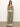 Against a plain background, a person wears "the soft trouser mezzo sage" by Sanctuary Clothing. The green sleeveless jumpsuit features an intricate floral and geometric pattern. They have long hair with sandals and necklaces, hands tucked in pockets.