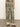 Someone wearing the Soft Trouser Mezzo Sage by Sanctuary Clothing, featuring an ornate green, beige, and brown pattern with a high-waisted, wide-leg design, paired with sandals against a plain light-colored wall.