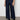 A person wears Sanctuary Clothing's the marine magnetic wide-leg, dark blue jeans with front patch pockets, paired with a black top and black ankle boots, standing against a light-colored wall and concrete floor.