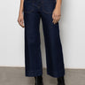A person wears Sanctuary Clothing's the marine magnetic wide-leg, dark blue jeans with front patch pockets, paired with a black top and black ankle boots, standing against a light-colored wall and concrete floor.