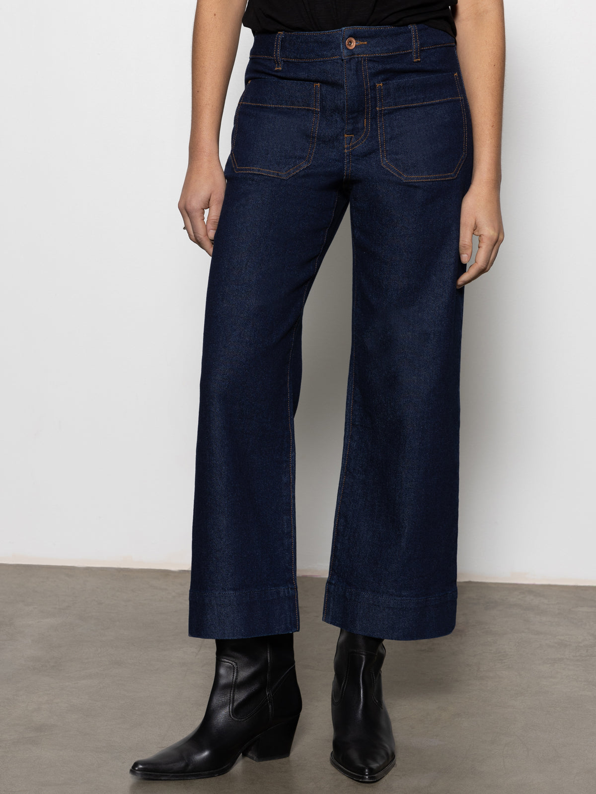 A person wears Sanctuary Clothing's the marine magnetic dark blue wide-leg jeans with front pockets, paired with a black top and black ankle boots, standing against a plain light background.