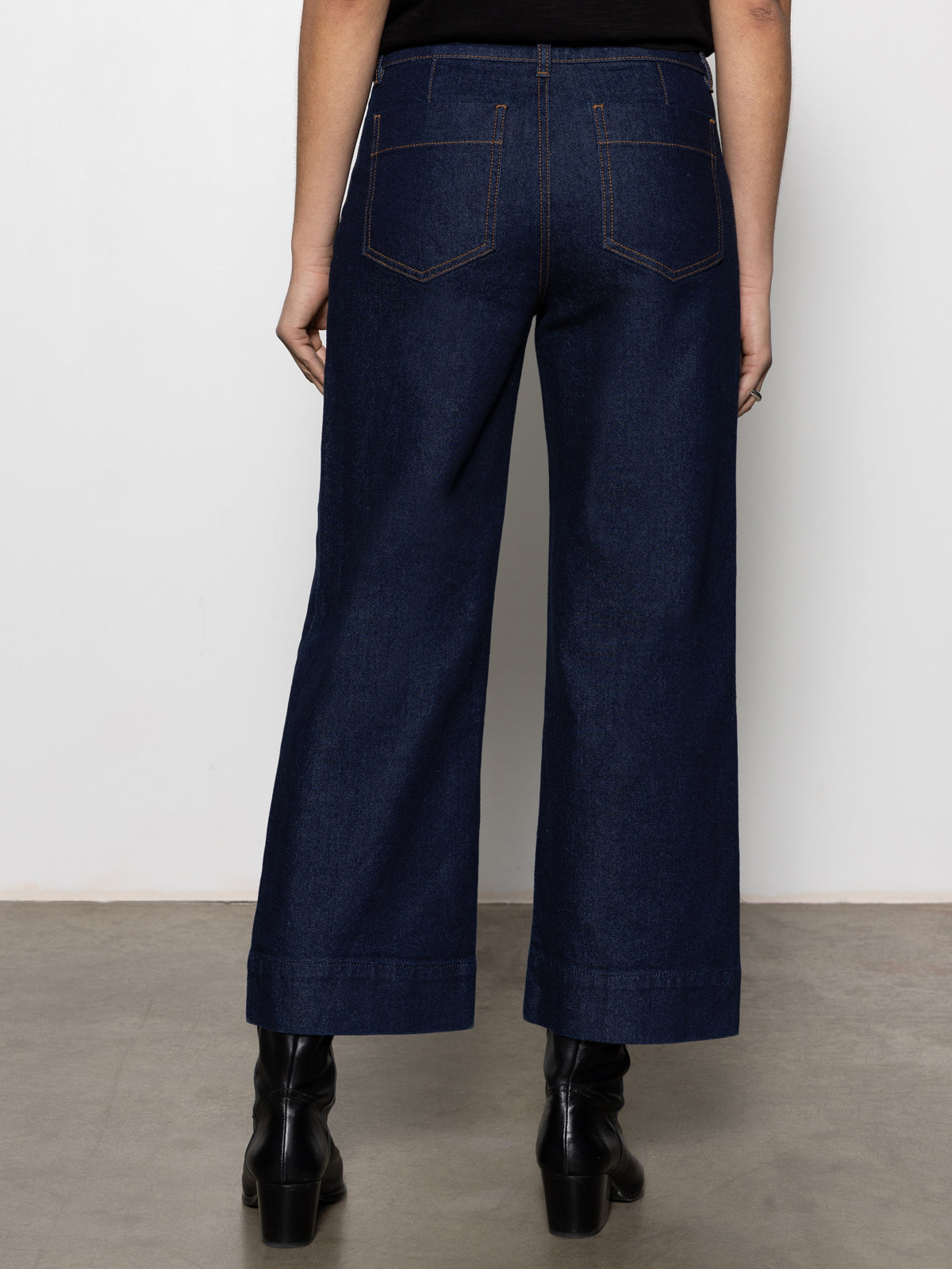 A person faces away, modeling Sanctuary Clothing's "the marine magnetic" dark blue wide-leg jeans with visible back pockets and black ankle boots, shown waist down against a plain background.