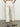A person is seen from behind wearing Sanctuary Clothing's "The Marine Crop Trouser Pant" in French Vanilla paired with a striped top. They stand in a minimalist room featuring a light gray wall, dark floor, and black and white patterned sandals.