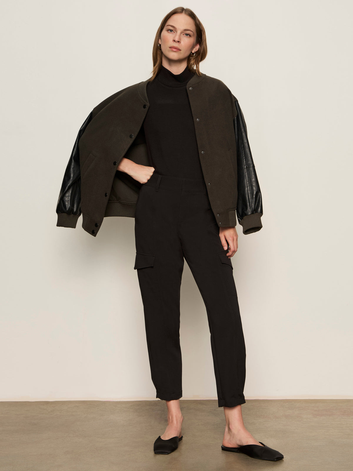 A woman stands indoors against a plain wall, wearing Sanctuary Clothingžs polished cargo pant in black, a dark bomber jacket draped over her shoulders, a black turtleneck, and black pointed-toe flats, looking at the camera with a neutral expression.