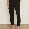 A person wears Sanctuary Clothing's polished cargo pant in black, featuring side pockets and a cropped fit, standing on a brown floor against an off-white wall with black slip-on shoes.
