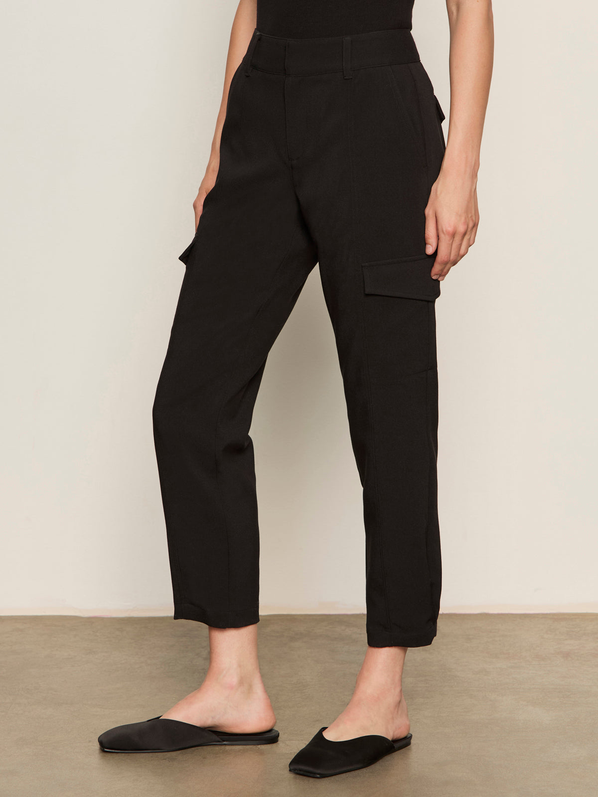 A person wearing the Sanctuary Clothing polished cargo pant in black and black slip-on shoes stands on a concrete floor against a light-colored wall.