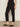 A person wearing the Sanctuary Clothing polished cargo pant in black and black slip-on shoes stands on a concrete floor against a light-colored wall.