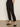 A person wears Sanctuary Clothing's polished cargo pant in black with black slip-on shoes, standing against a plain light background and posing with one hand in their pocket.