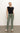 A person wearing a black sleeveless top and Sanctuary Clothing's sculpted Hayden bootcut pants in dark spruce stands casually with one hand in their pocket. They sport white shoes with black accents against a plain, neutral background.