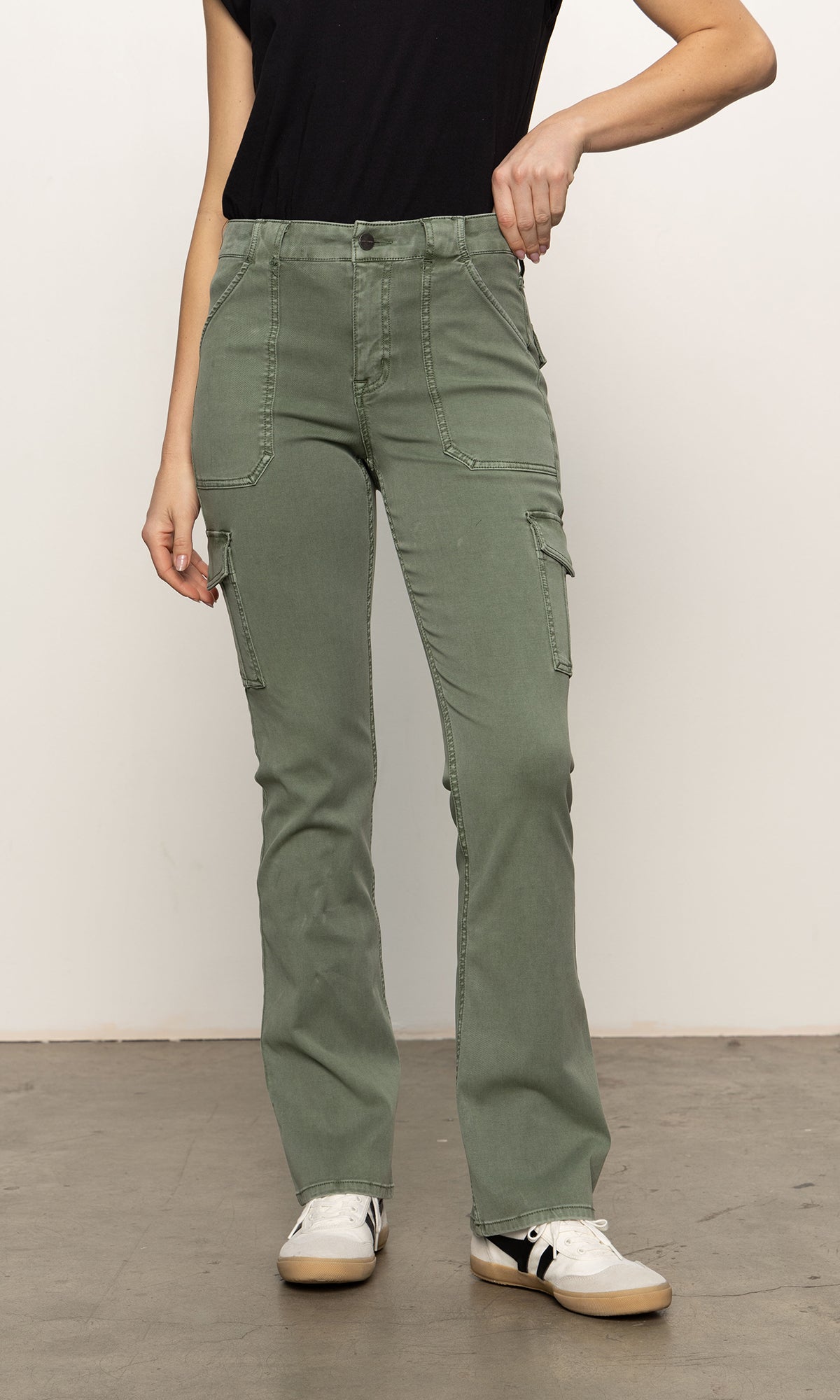 A person in a black T-shirt and Sanctuary Clothing's sculpted Hayden bootcut pant in dark spruce stands on a concrete floor. They rest one hand on their hip, highlighting their casual white sneakers with black details.