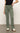 A person in a black T-shirt and Sanctuary Clothing's sculpted Hayden bootcut pant in dark spruce stands on a concrete floor. They rest one hand on their hip, highlighting their casual white sneakers with black details.