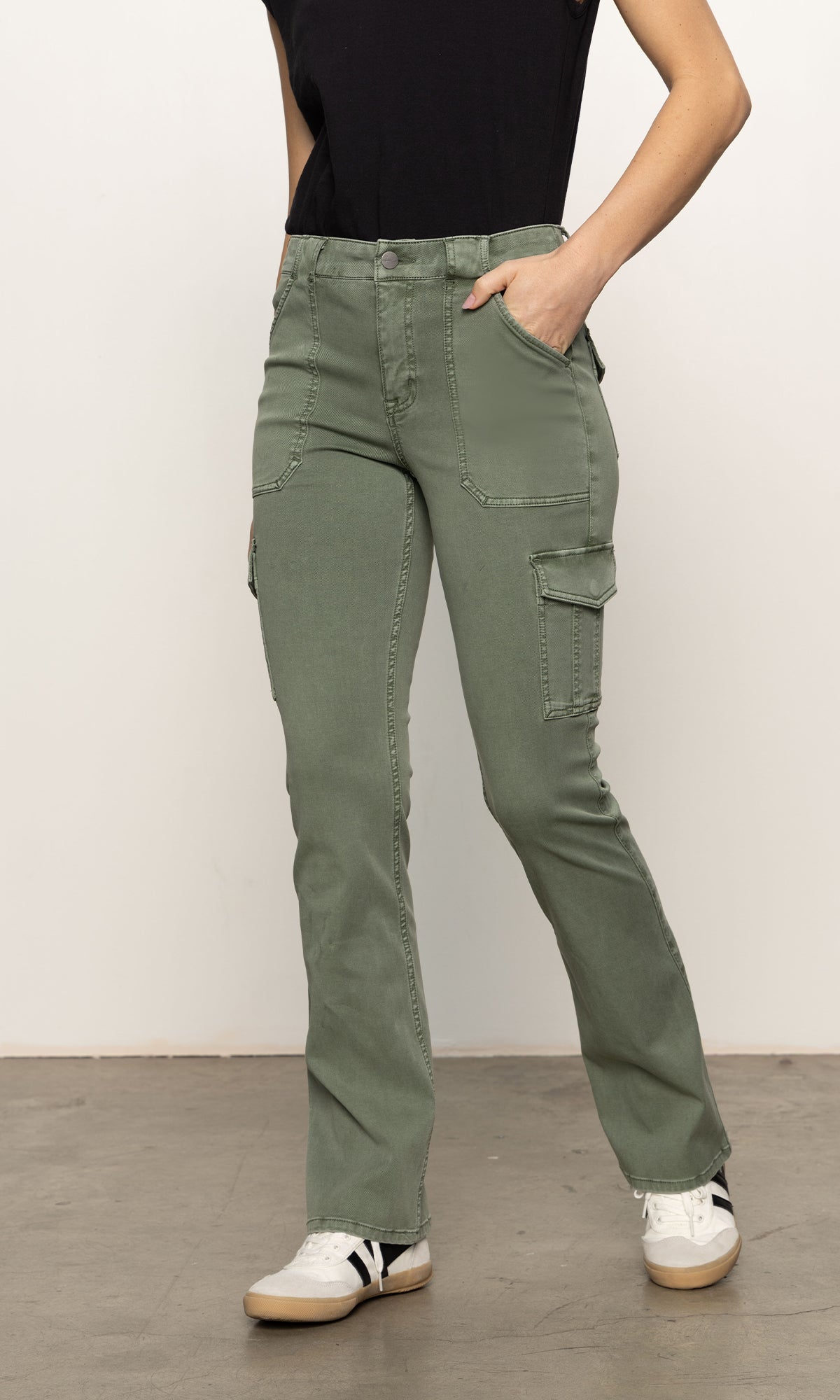 A person dressed in Sanctuary Clothing's "sculpted hayden bootcut pant dark spruce," paired with a black top and white sneakers, casually stands on a concrete floor with one hand in their pocket against a plain white background.