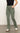 A person dressed in Sanctuary Clothing's "sculpted hayden bootcut pant dark spruce," paired with a black top and white sneakers, casually stands on a concrete floor with one hand in their pocket against a plain white background.