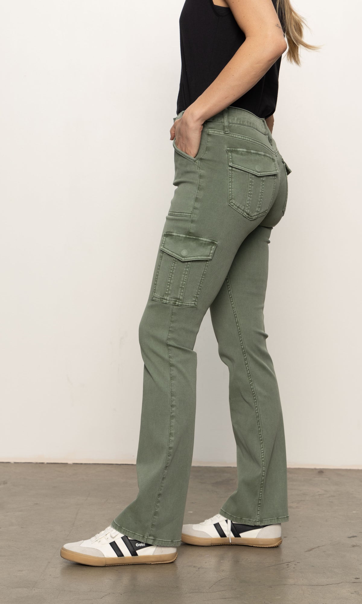 A person in Sanctuary Clothing's sculpted Hayden bootcut pant in dark spruce and a black sleeveless top stands in profile. The pants feature side and back pockets. They're also wearing white sneakers with black stripes against a simple white wall and gray flooring.