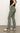 A person in Sanctuary Clothing's sculpted Hayden bootcut pant in dark spruce and a black sleeveless top stands in profile. The pants feature side and back pockets. They're also wearing white sneakers with black stripes against a simple white wall and gray flooring.