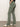 Someone in dark spruce sculpted Hayden bootcut pants by Sanctuary Clothing and white sneakers with black stripes stands against a plain wall. The pants include side and back pockets, and the person wears a black top. The floor is concrete.
