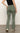A person in Sanctuary Clothing's sculpted Hayden bootcut pants in dark spruce and a black top stands on a plain floor against a white background, with one hand on their hip and wearing white sneakers featuring black accents.