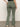 A person wearing Sanctuary Clothing's "sculpted hayden bootcut pant" in dark spruce is standing against a plain background with their left hand on their hip. The pants, featuring multiple pockets, are paired with white sneakers.