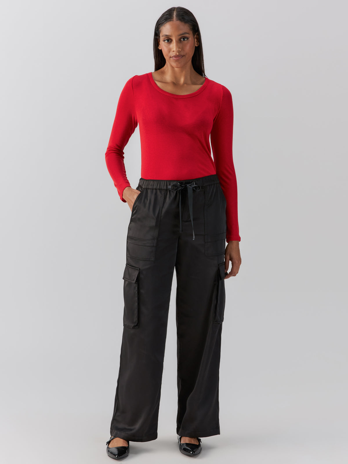 A person stands against a plain background wearing a long-sleeved red top and black Satin Cargo Wide Leg pants from Sanctuary Clothing with hands in pockets. They have long, dark hair and are wearing black flats.