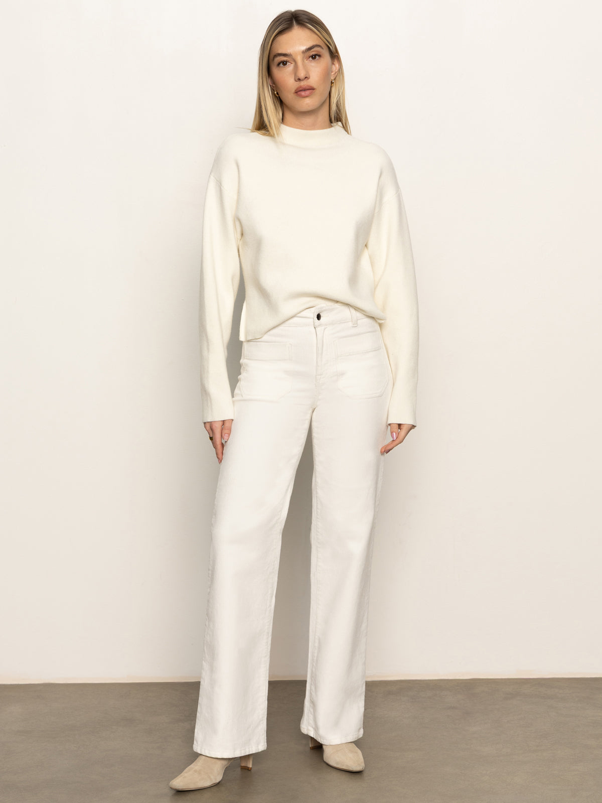 A woman with straight blonde hair and a neutral expression stands against a plain background, wearing Sanctuary Clothing's corduroy marine pant in chalk, paired with a cream sweater and beige shoes.