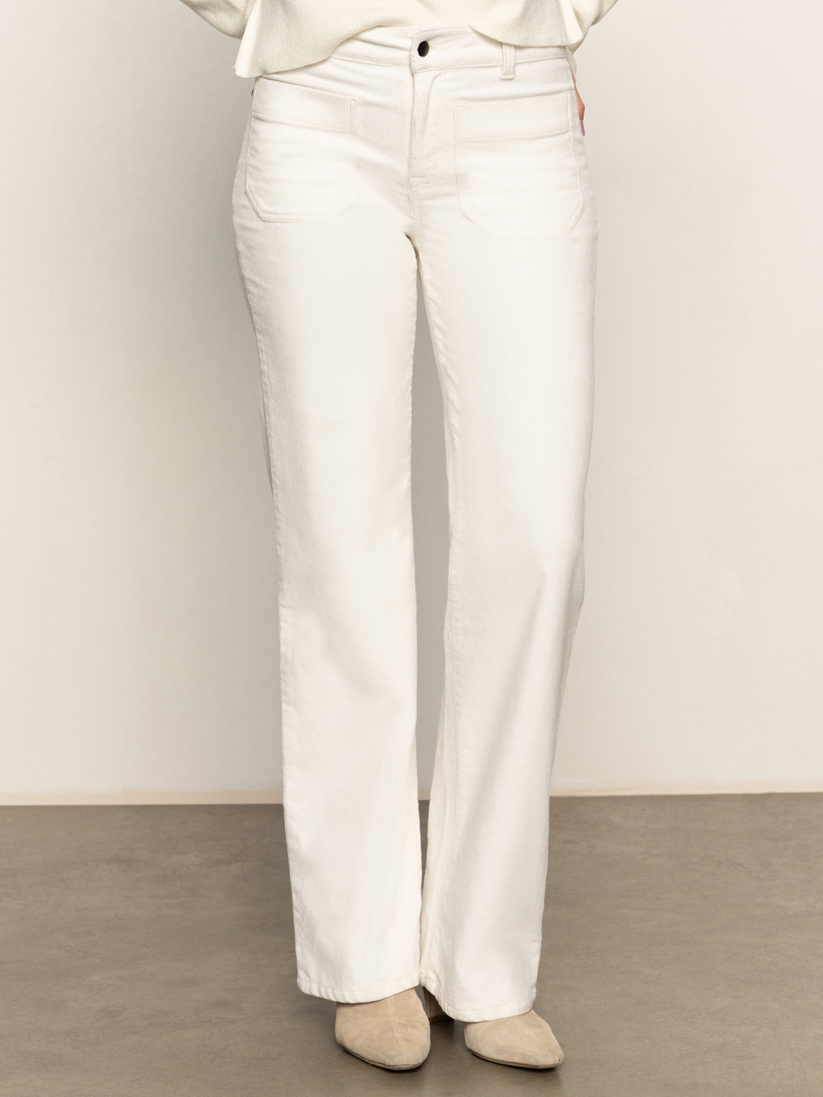 A person wears Sanctuary Clothing's corduroy marine pant in chalk, paired with light beige suede shoes, standing on a gray floor against a neutral background.