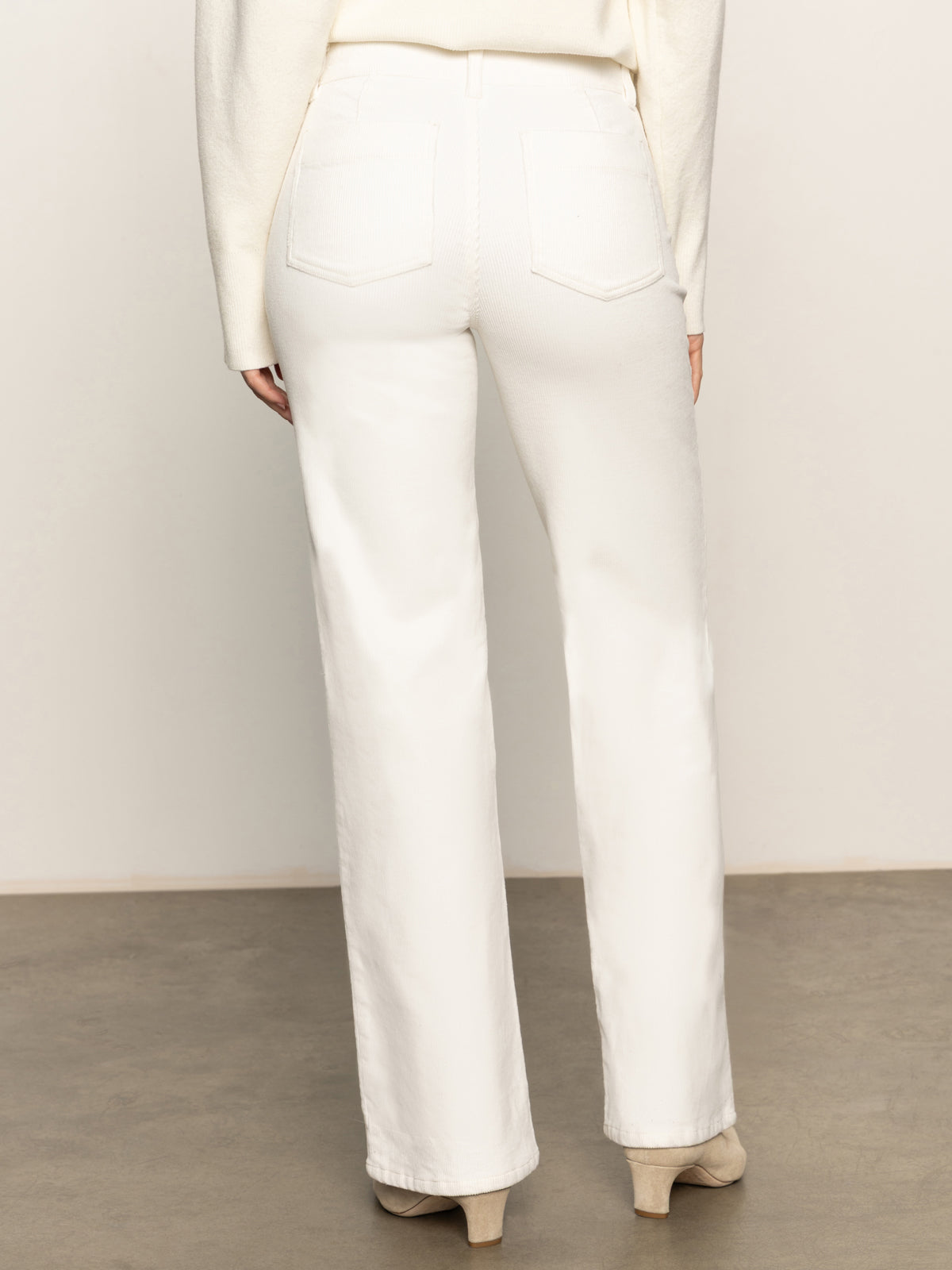 Back view of a person wearing Sanctuary Clothing’s corduroy marine pant in chalk with patch pockets, paired with a cream sweater and beige heeled boots, standing on a gray floor against a neutral background.