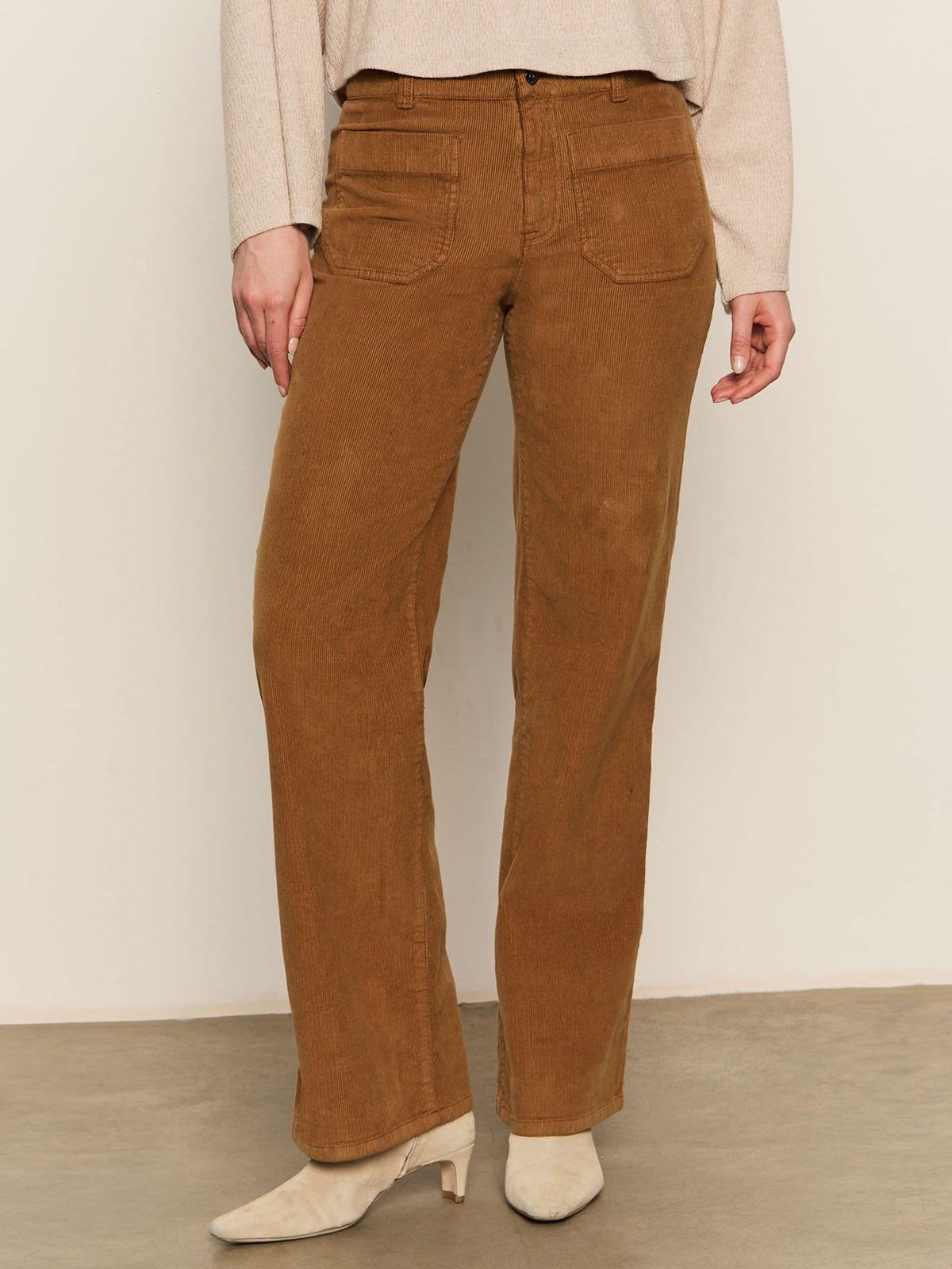 Someone wearing Sanctuary Clothing's corduroy marine pant in bronze, paired with a light beige long-sleeve top and cream heeled boots, stands against a plain background.
