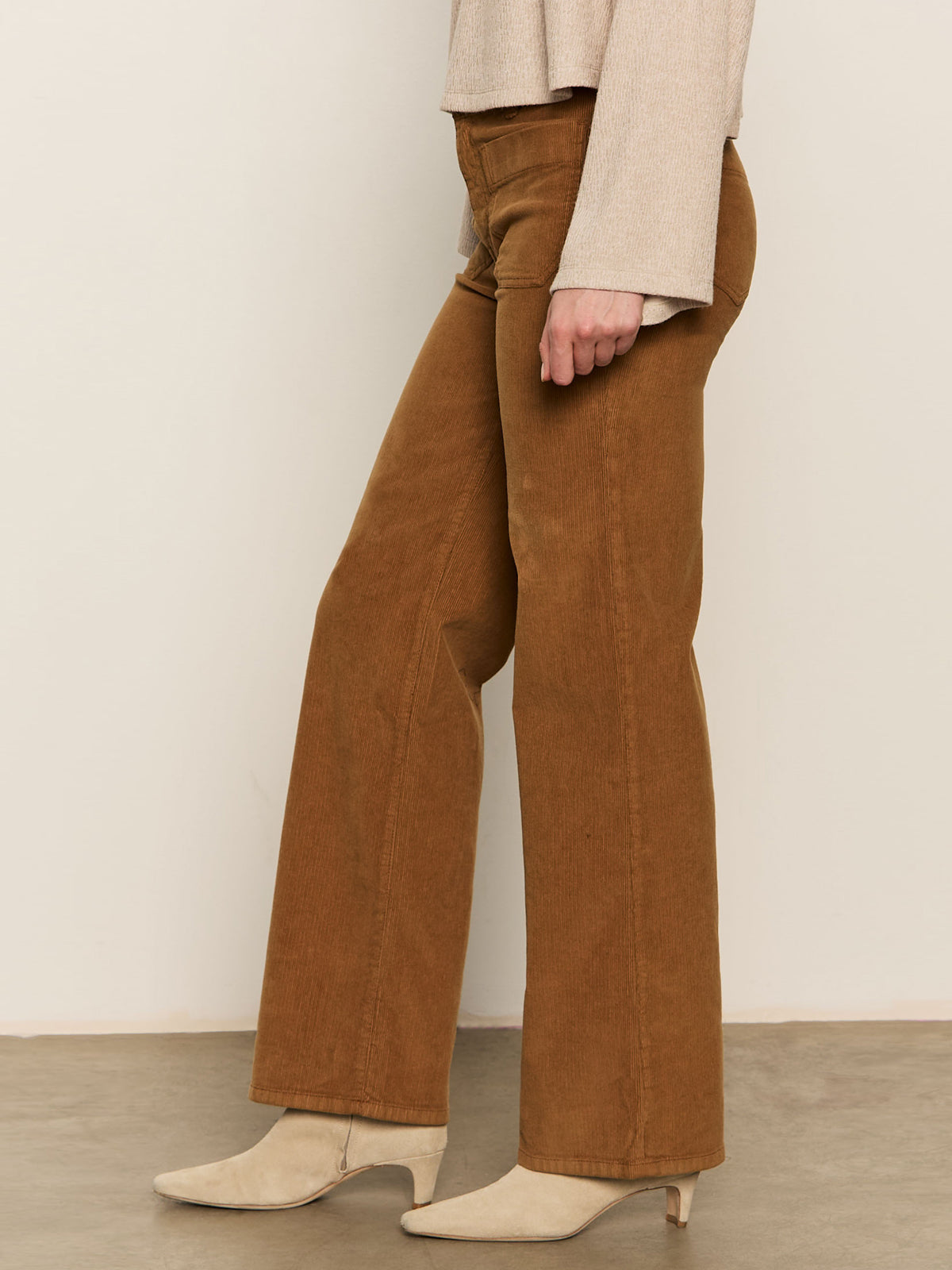 Someone stands on a smooth concrete floor against a plain light wall wearing Sanctuary Clothing's corduroy marine pant in bronze, paired with a beige long-sleeve top and cream heeled ankle boots.