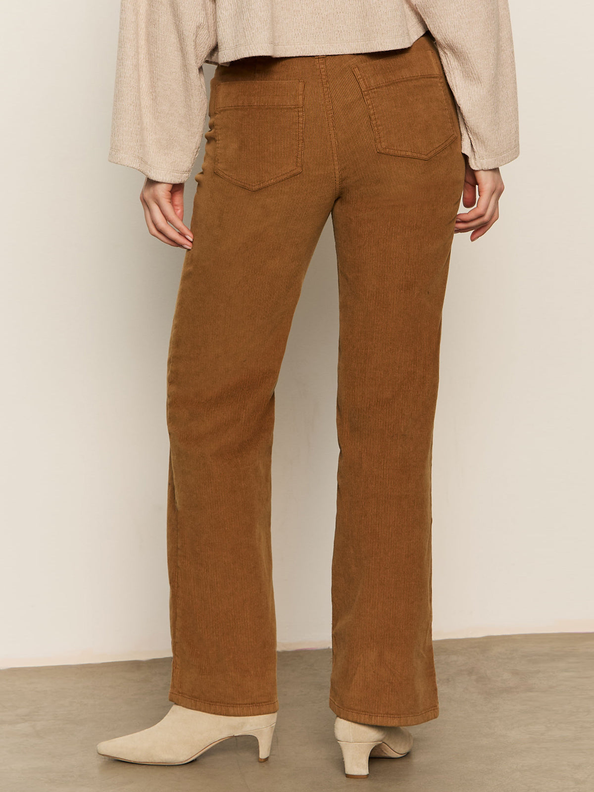 A person faces away from the camera against a neutral background, wearing Sanctuary Clothing’s corduroy marine pant in bronze, paired with a beige long-sleeve top and cream heeled boots.