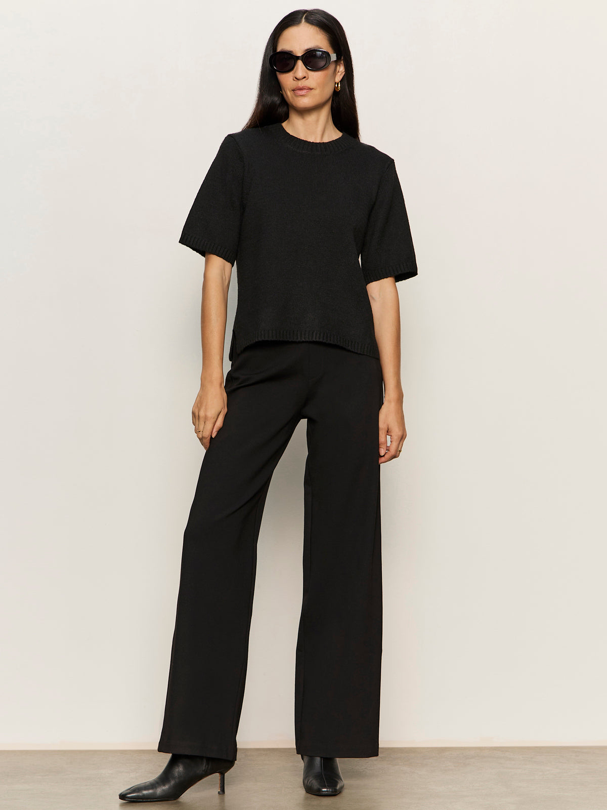 A woman stands against a light background wearing sunglasses, a black short-sleeve sweater, and black heeled boots, paired with the runway ponte trouser in black by Sanctuary Clothing.