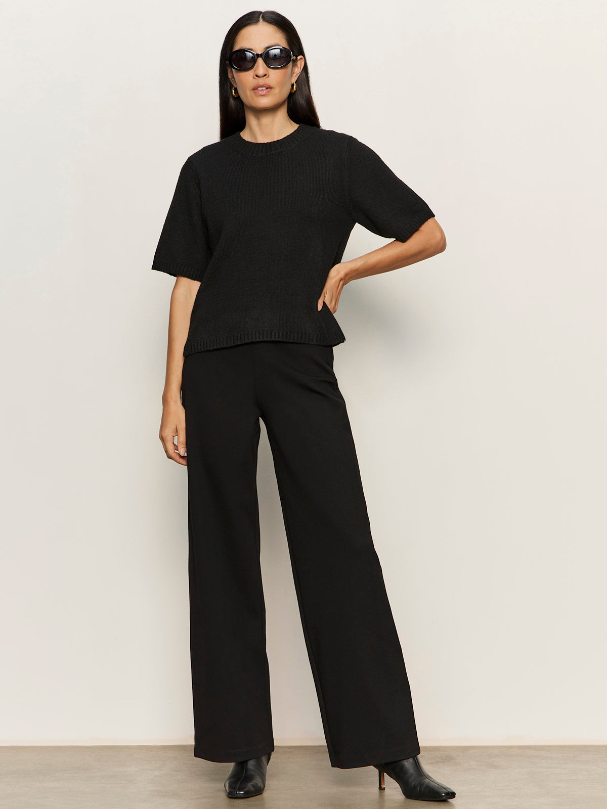 A woman stands confidently against a plain light background, wearing black sunglasses and Sanctuary Clothing’s runway ponte trouser in black, paired with a short-sleeved black top and heeled ankle boots. One hand rests on her hip, the other by her side.