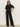 A woman stands confidently against a plain light background, wearing black sunglasses and Sanctuary Clothing’s runway ponte trouser in black, paired with a short-sleeved black top and heeled ankle boots. One hand rests on her hip, the other by her side.