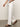 A person in Sanctuary Clothing's white arden wide-leg jeans and brown sandals walks on a concrete floor, cropped above the waist, with one hand visible holding a shirt.