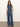 A woman with long brown hair stands against a plain background wearing Sanctuary Clothing’s Arden Wide Leg Jeans in Perfect Storm and a sleeveless, button-up denim top. She completes the look with sandals and one hand in her pocket.