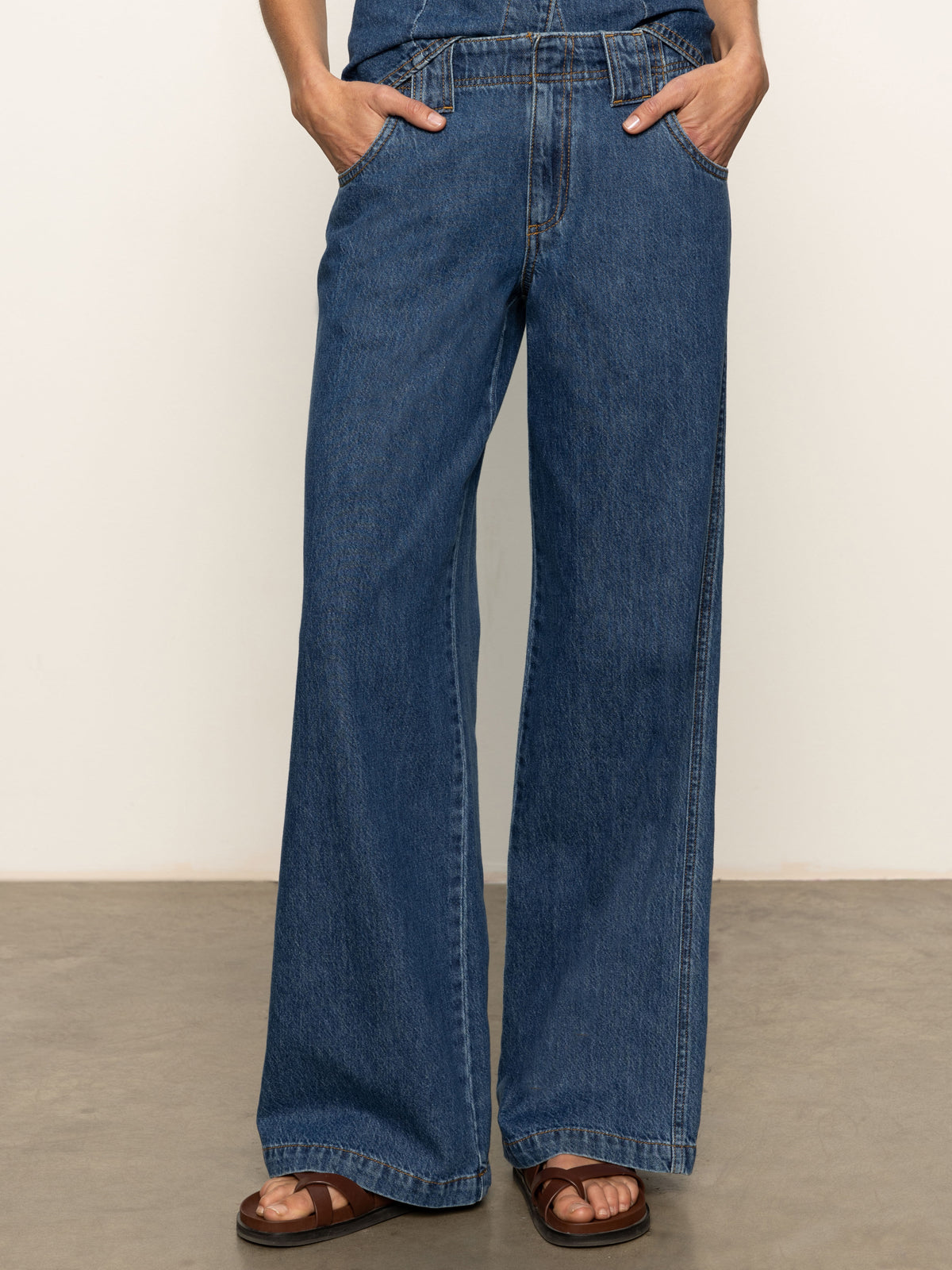 A person models Sanctuary Clothing's arden wide leg jeans in the shade perfect storm, standing with hands in pockets and paired with brown sandals against a plain background.