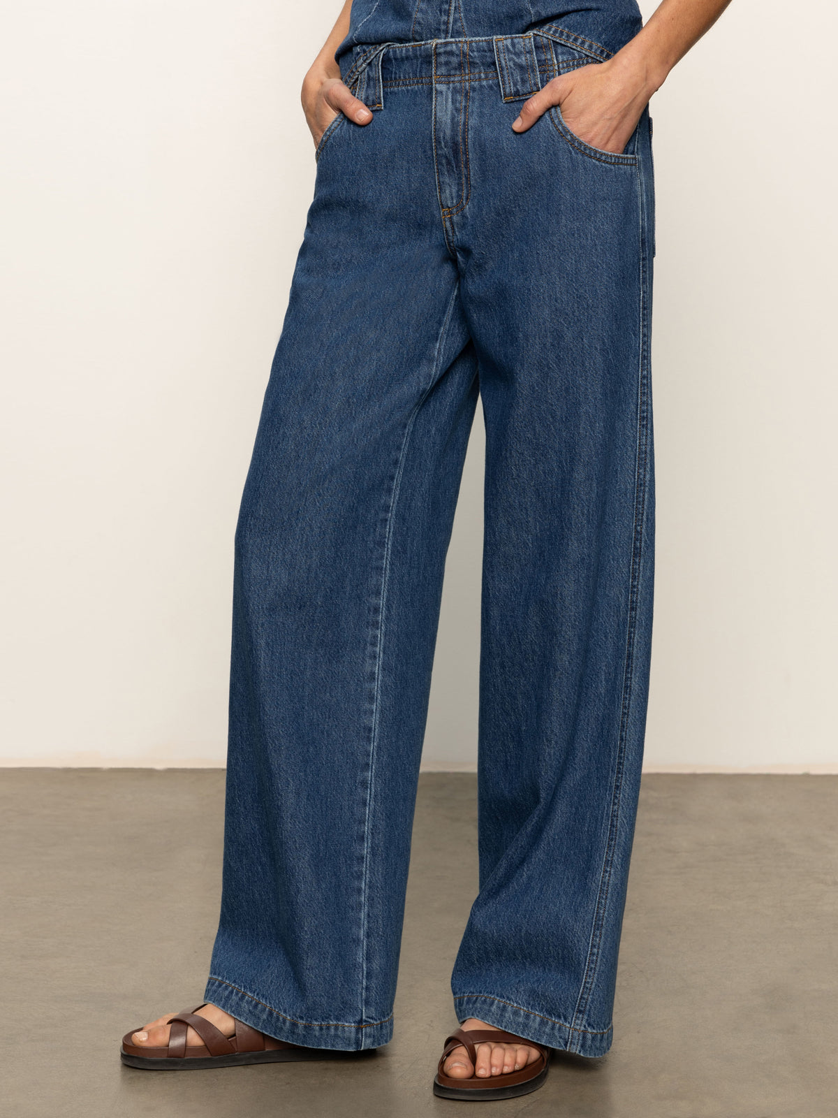A person stands with hands in pockets, wearing Sanctuary Clothing’s arden wide leg jeans perfect storm and brown flat sandals against a plain light background.