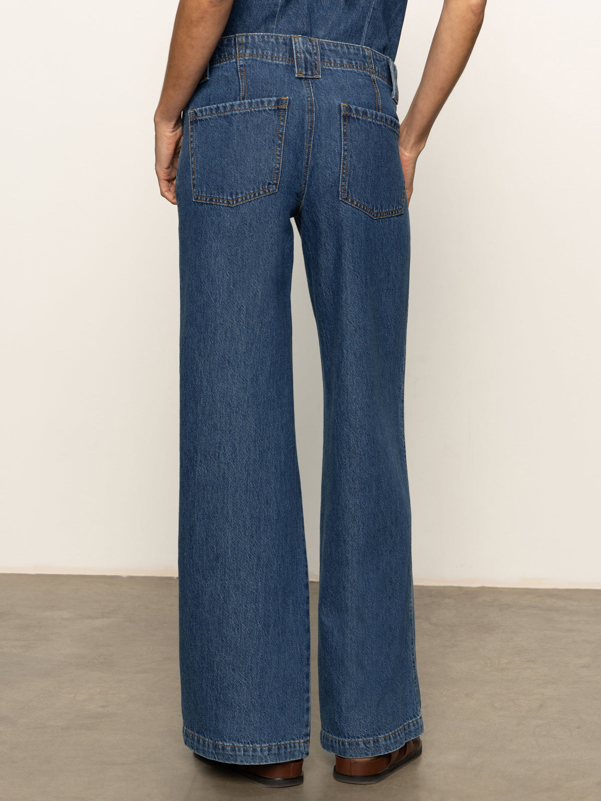 A person wearing Sanctuary Clothing's arden wide leg jeans in Perfect Storm stands on a polished concrete floor, facing away to show the two large back pockets.