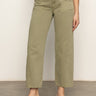 A person wearing Sanctuary Clothing's district column ankle jean bamboo—high-waisted, olive green wide-leg jeans with front patch pockets—paired with a white top and black heeled sandals, stands on a gray floor against a light background.