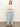 A woman stands against a plain background wearing Sanctuary Clothing’s district column ankle jean in blue mirage, paired with a white long-sleeve top and brown sandals. She has long, straight hair and a neutral expression, with one hand on her thigh.