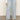 A person wears Sanctuary Clothing’s district column ankle jean in blue mirage with front pockets, a white long-sleeve top, and brown sandals, standing on a gray floor against a plain light background.
