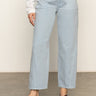 A person wears Sanctuary Clothing’s district column ankle jean in blue mirage with front pockets, a white long-sleeve top, and brown sandals, standing on a gray floor against a plain light background.
